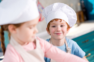 adorable smiling little boy in chef hat looking at friend cooking on foreground