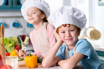 adorable siblings in chef hats and aprons smiling at camera while cooking in kitchen