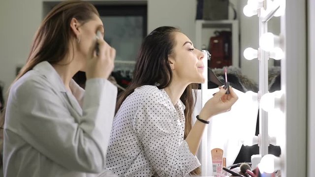 Beautiful women friends doing makeup standing before mirror