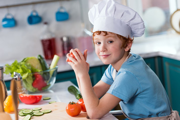 cute little boy in chef hat smiling at camera while cooking in kitchen