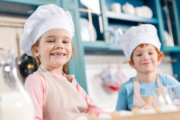 adorable children in chef hats and aprons smiling at camera while cooking together in kitchen