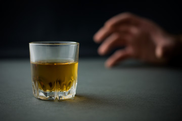 hand reaches for a glass of whiskey or cognac or alcohol drink, alcoholism and alcohol abuse concept, defocused, selective focus, close up, gray table, dark background