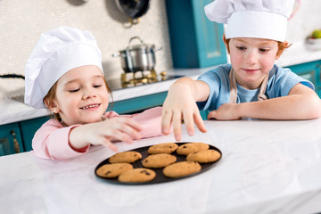 happy little kids in chef hats eating tasty cookies in kitchen
