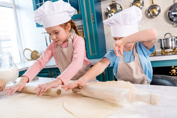 focused little children in chef hats and aprons rolling dough in kitchen