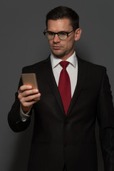 Serious thoughtful businessman with glasses and formal suit looking at the smartphone screen on gray background