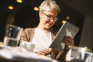 Pretty girl with glasses and short hair checking the news on a tablet while drinking a coffee.