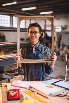 Close up vertical view of cheerful happy middle aged engineer holding old fashion picture frame in sunny fabric workshop and posing behind the desk with tools.