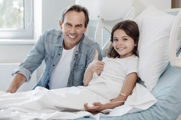 Fototapeta premium Everything is cool. Charming young lady thumbing up and smiling while lying in a hospital bed and getting support from her loving daddy.