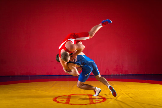 Two young men wrestlers in blue and red wrestling tights are wrestlng and making a hip throw on a yellow wrestling carpet in the gym, sied view