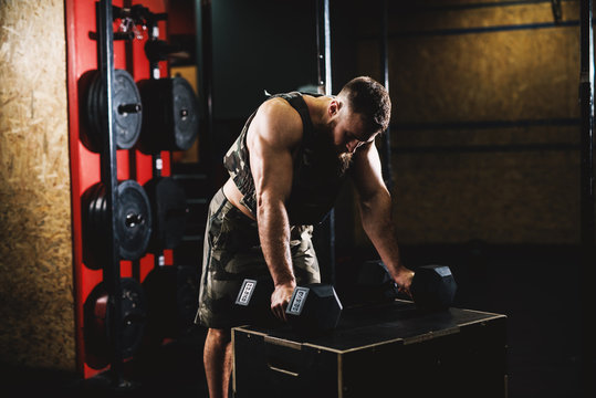 Sporty shape bodybuilder guy in military vest holding dumbbells on the large wooden box and preparing for exercise in the dark gym.