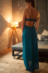 Young woman in blue dress stands against background of bed and lamp. Back view.