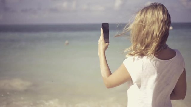 young, beautiful woman using her phone at the beach and taking pictures