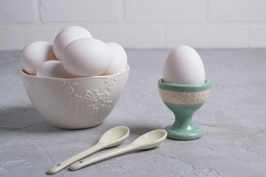 White  chicken  egg in ceramic stand and eggs in white  bowl . On a gray concrete background.