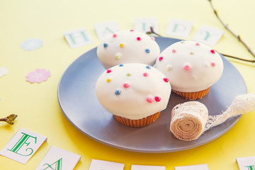 Plate with delicious Easter cupcakes and vintage ribbon with Happy Easter lettering, fresh branches with kidneys and paper flowers on bright yellow background. Art concept. Selective focus.