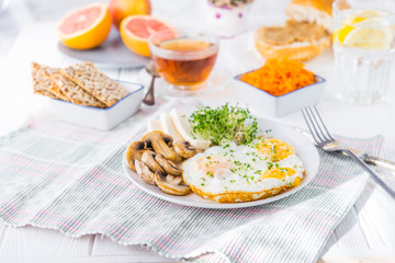 Healthy breakfast plate with scrambled eggs, cheese, grilled mushrooms and sprout micro greens and other snacks and drinks on the served white wooden table. Selective focus.