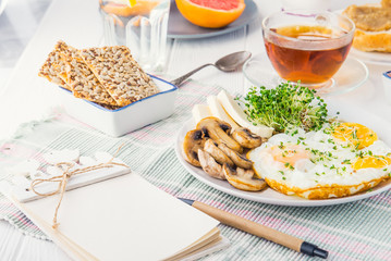 Notebook with emty blanks and healthy breakfast plate with scrambled eggs, cheese, grilled mushrooms and sprout micro greens on the served white wooden table. Selective focus.