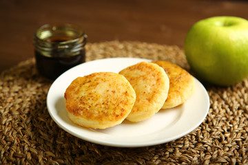 Cheese pancake from cottage cheese are on a white plate. Nearby there is a glass jar with honey and an apple. Wooden background, woven backing. Proper nutrition. Dark background