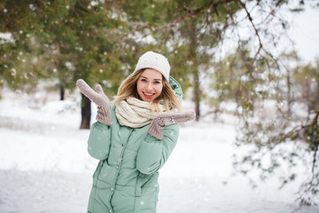 A cute blonde girl outdoors in winter, smiling and looking at the camera, it's snowing. Young beautiful girl raised her hands up, laughs and catches snowflakes. Winter theme. Space for text.