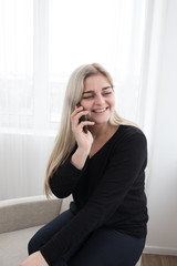 white blonde girl sitting on a white armchairon a white background in black pullover and posing and smiling with her smartphone talking with friends