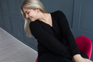 white blonde girl sitting on a red eiffel chair on a grey dark grey background in black pullover...