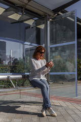 Business woman in casual clothes carefully reviews something important on her mobile phone, while waiting sitting at the bus stop, on a sunny day