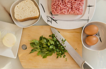 ingredients for cooking meat cutlets on the table, pork minced meat on a plate, onion, eggs, glass of milk, sliced parsley, knife, cutting kitchen Board, bread