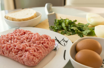 ingredients for cooking meat cutlets on the table, pork minced meat on a plate, onion, eggs, glass of milk, sliced parsley, knife, cutting kitchen Board, bread, cooking