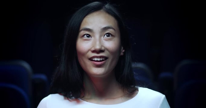Close up, slow motion shot of an asian woman in a movie theatre reacting in awe.