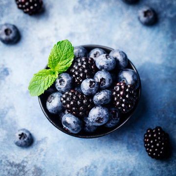 Blueberry and blackberry berries with mint leaves in black bowl on blue stone background
