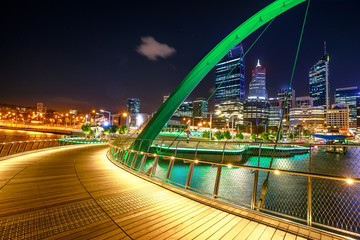 Wooden footpath of Elizabeth Quay Bridge illuminated by night at entrance of Elizabeth Quay marina, a new tourist attraction in Perth, Western Australia.Esplanade with modern skyscrapers on background