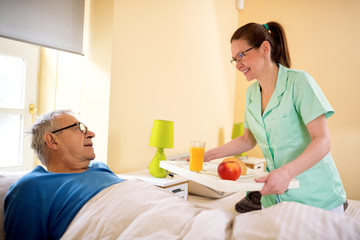 Young smiling nurse brings breakfast to senior patient
