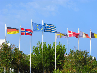 Flags of EU, Greece, Germany, Norway, Sweden, Denmark, France above green trees in blue sky.