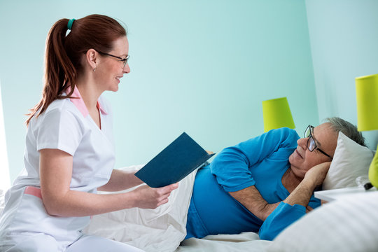 Smiling positive nurse reading a book to her old patient