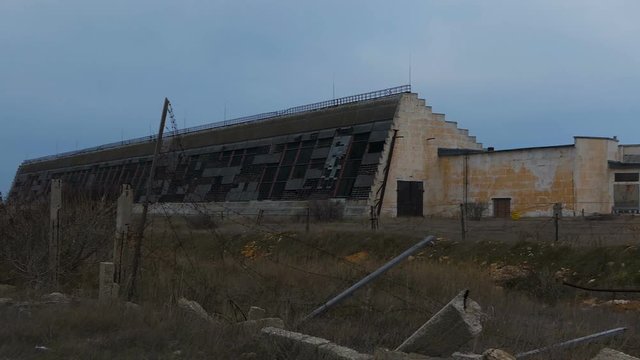 Abandoned military hangars and buildings in a closed area with barbed wire. Technical territory with technical objects. The camera moves slowly along the buildings.