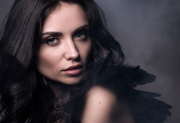 Black angel in a cloud of smoke. Beauty closeup portrait in dark tones. Sexy young woman in black with black feathers