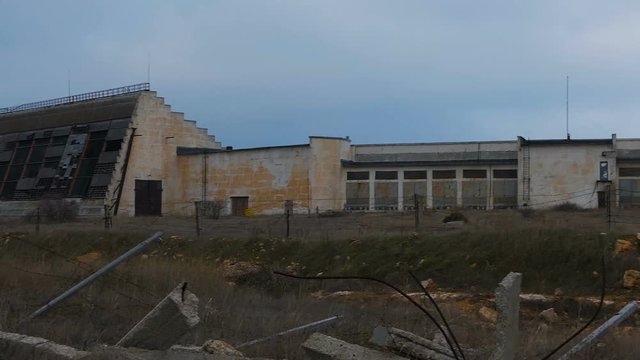 Abandoned military hangars and buildings in a closed area with barbed wire. Technical territory with technical objects. The camera moves slowly along the buildings.
