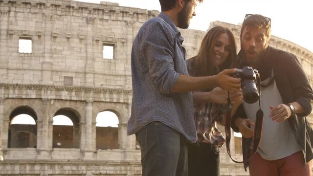 Three young friends tourists in front of colosseum in rome at sunset have fun looking browsing pictures photos on dslr camera happy beautiful girl long hair slow motion