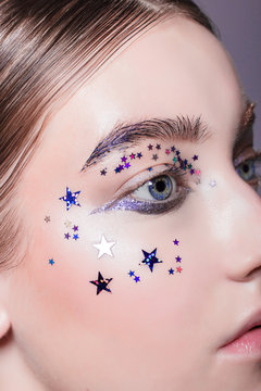 The beautiful luxury brunette with dense eyebrows and eyelashes show a fashionable festive make-up. Gloss, sequins, stars. Close up of a girl's face with open eyes