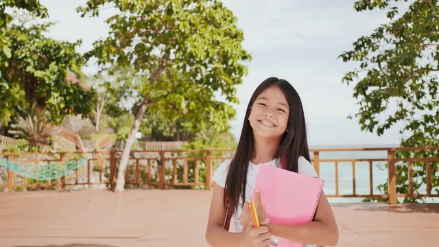 A charming philippine schoolgirl with a backpack and books in a park off the coast. A girl joyfully poses, raising her hands up with textbooks in her hands. Warm sunny day.