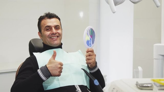 Cheerful handsome mature male patient sitting in the dental chair looking in the mirror smiling showing thumbs up curing cavity treatment health medicine dentistry clinical examination.