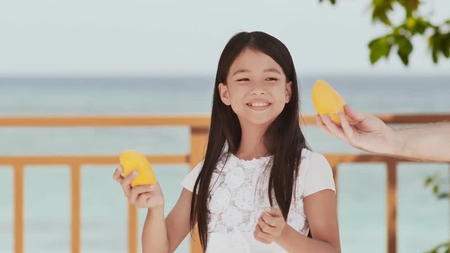 A charming filipino girl schoolgirl in a white dress and long hair takes a mango from a man's hand. The sun. The blue ocean. Childhood. Recreation.