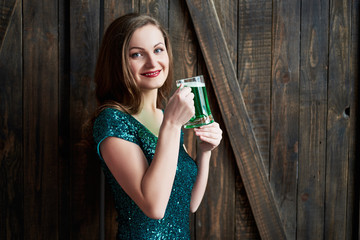 Obraz premium Sexy smiling woman holding mug of green beer for St. Patricks Day celebrations on wooden background, copy space. Girl in emerald sequins dress holding glass of beer in pub. Greeting card, March 17