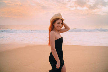 Fototapeta na wymiar young red-haired woman walking along the beach at sunset, smiling and looking at the camera