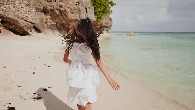 A charming and happy philippine teenage girl in a white summer dress is running along a tropical beach near the rocks. She is happily spinning. Childhood. Recreation.