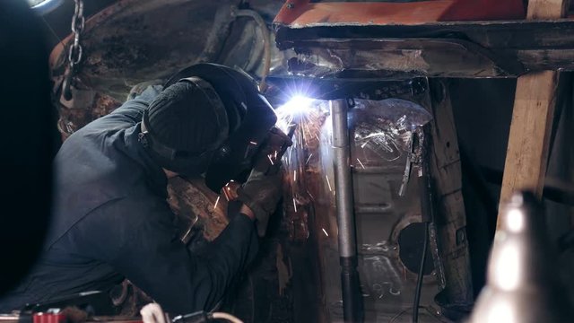Backside footage of a welder joining some metal pieces together on car's bottom surface with a welding torch.