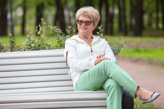 Smiling European Elderly Woman Sitting On The Edge Of White Bench In The Park