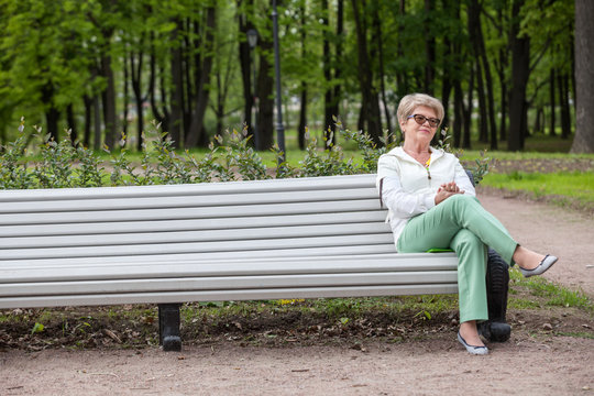 European Elderly Woman Sitting On The Edge Of Bench In The Summer Park, Copy Space