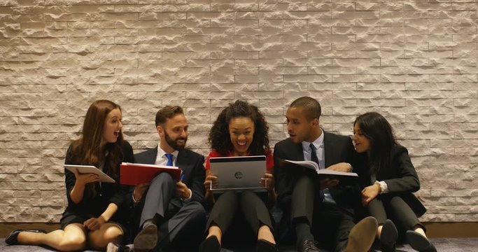 4K Relaxed business team in office, sitting on the floor using various computer devices & reacting to something exciting seen on computer screen