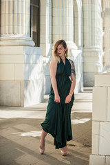 Alternative blonde model standing among white stone columns in emerald green halter top gown.