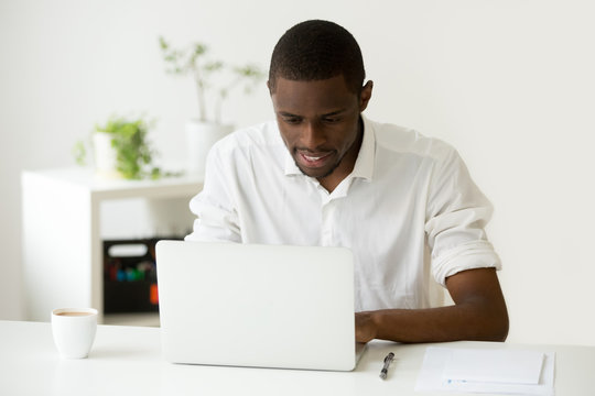 Smiling african man using laptop sitting at home office desk, black businessman or entrepreneur working on computer looking at screen, browsing web, search new job online, chatting in social networks